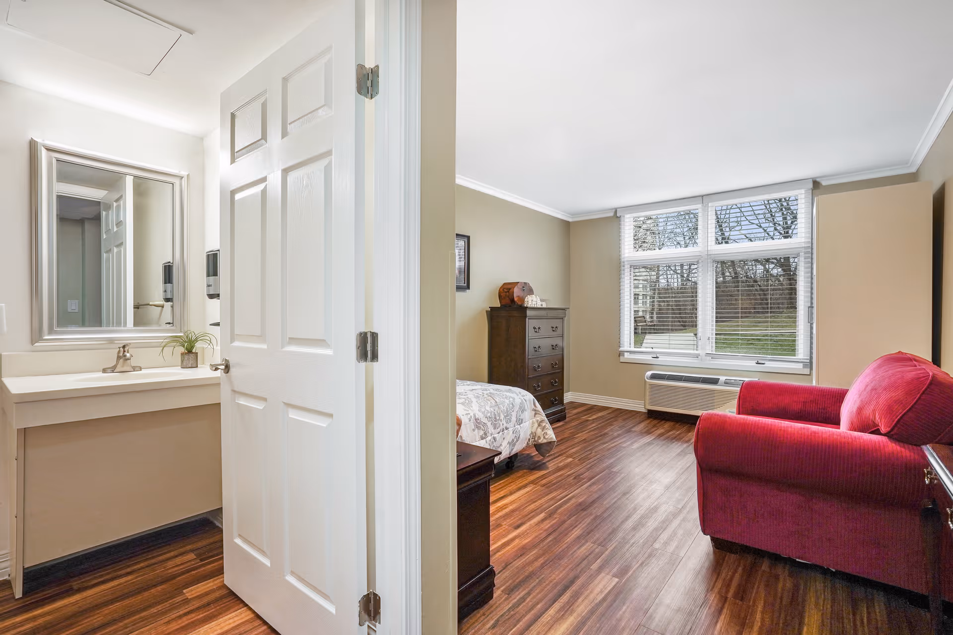 A bright senior living facility room with a wooden floor, a large window with blinds, a red armchair, a wooden dresser, and a bed partially visible. To the left, there is a small bathroom area with a white sink, a mirror, and a small plant on the counter.