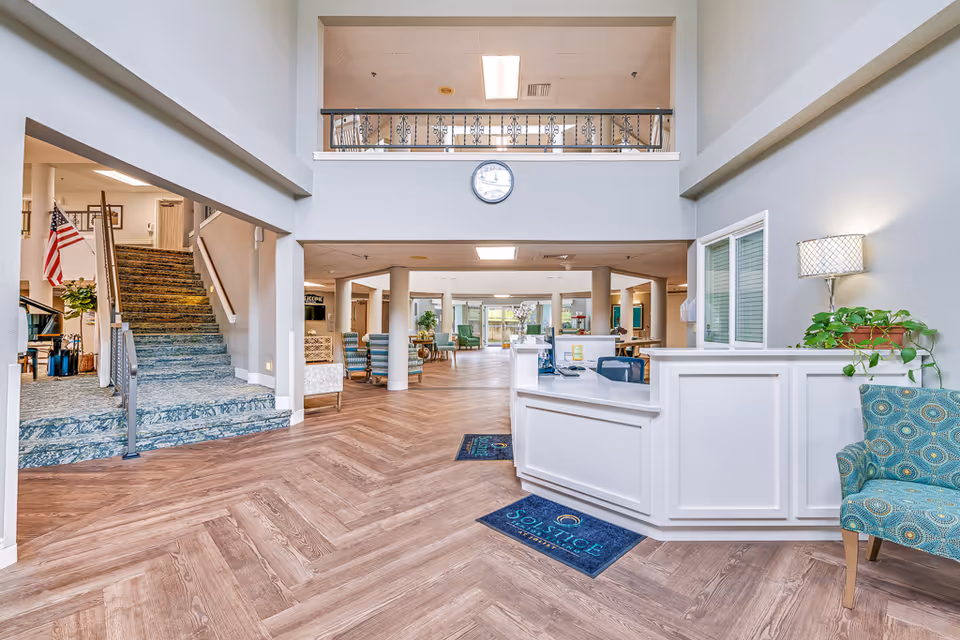Bright two-story reception lobby with a white front desk, seating areas, a staircase, and herringbone wood flooring.
