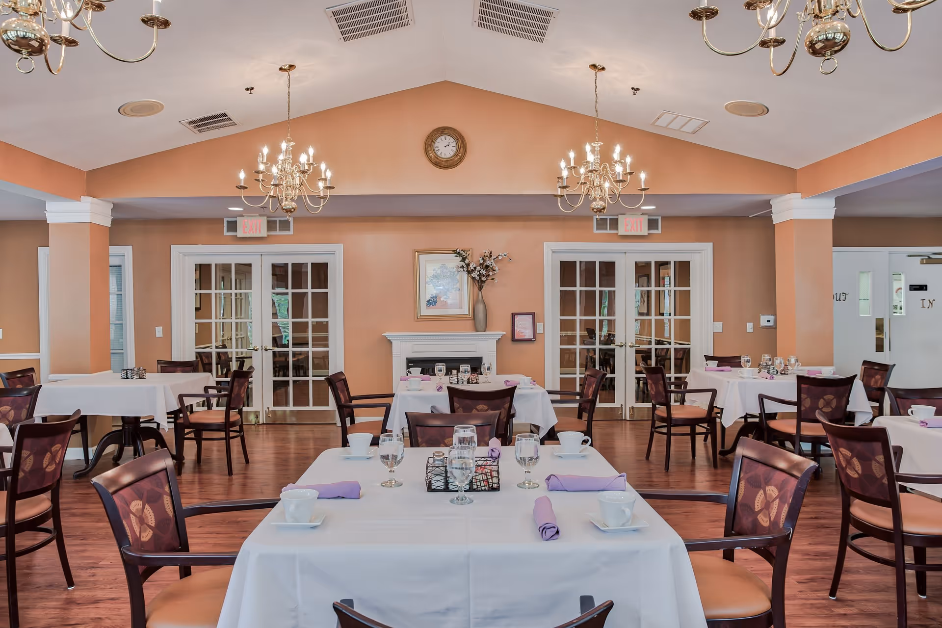 A dining room with several tables covered in white tablecloths, each set with cups, glasses, and purple napkins. The room has wooden chairs with patterned backs, a wooden floor, peach-colored walls, two chandeliers, and a clock above a decorative fireplace. French doors and exit signs are visible in the background.