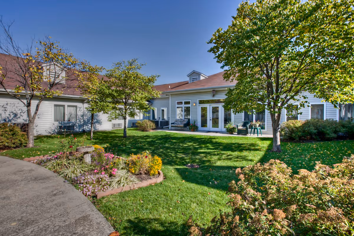 A sunny outdoor garden area at Bloom at German Church featuring a well-maintained lawn, flower beds with colorful flowers, several trees, and a light gray building with white-framed windows and doors in the background. There are outdoor chairs and a bench near the building, and the sky is clear and blue.