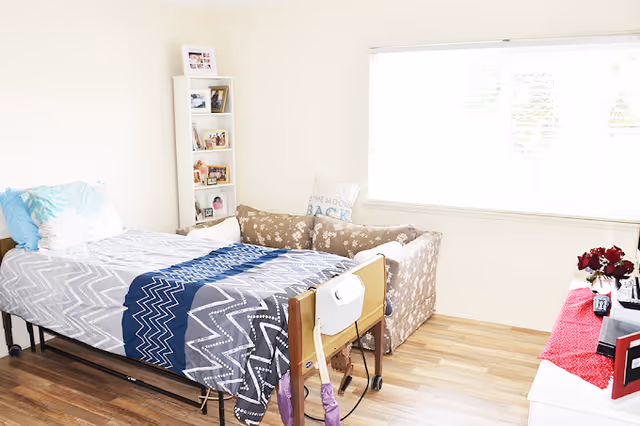 Bright resident bedroom with a medical single bed, patterned couch, small shelving unit, and a large window.