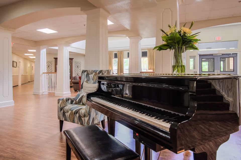 Interior view of a senior living facility common area featuring a black grand piano with a vase of flowers on top, a patterned armchair, wooden flooring, white columns, and large windows letting in natural light.