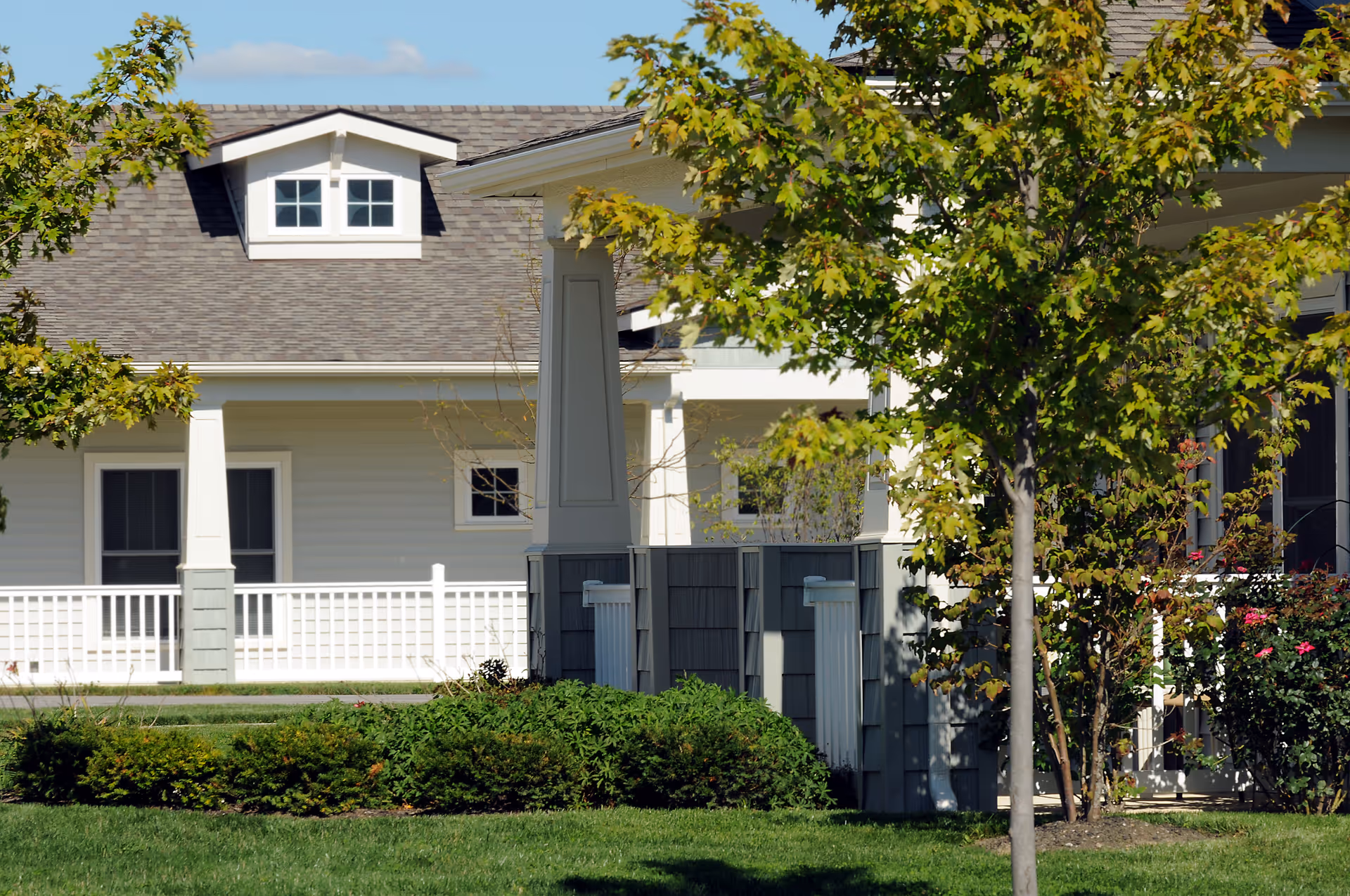 Front exterior of a light-colored residential building with a porch, white railing and columns, framed by trees and shrubs.