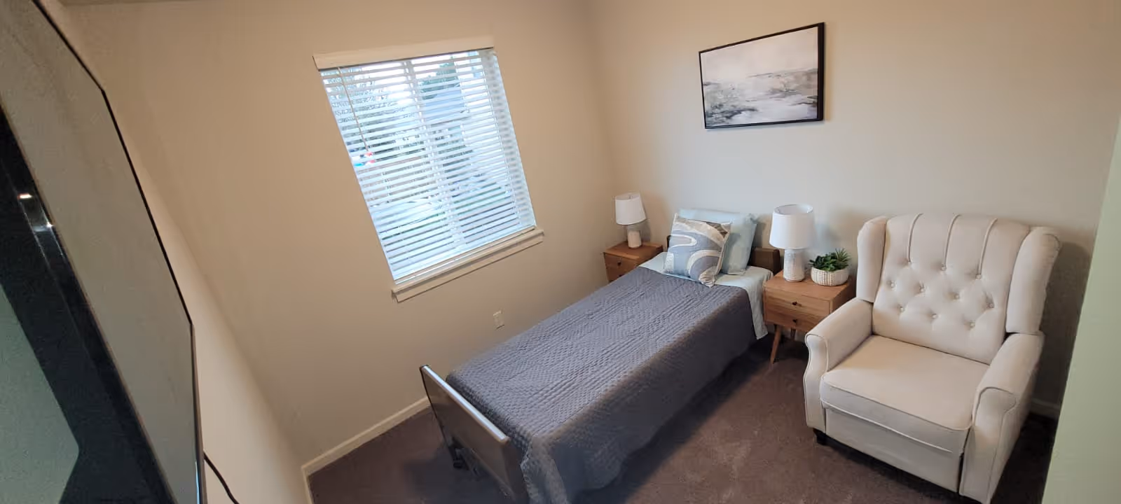 A small bedroom with a single bed covered in a gray quilt, two pillows, and two wooden nightstands with lamps and a small plant. There is a white upholstered armchair with button tufting next to the bed. A window with white blinds is on the left wall, and a framed abstract artwork hangs above the bed.
