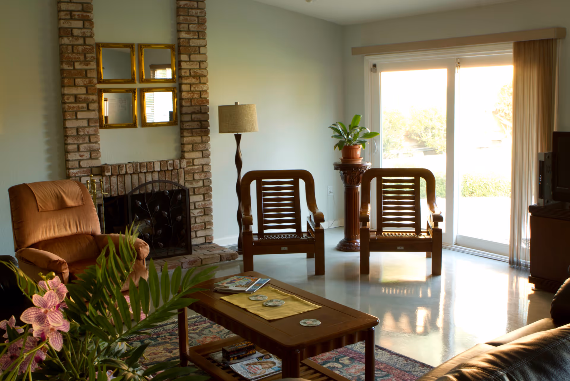 A cozy living room with a brick fireplace, a brown recliner chair, two wooden armchairs, a wooden coffee table with magazines and coasters, a floor lamp, a potted plant on a decorative pedestal, and a sliding glass door letting in natural light.