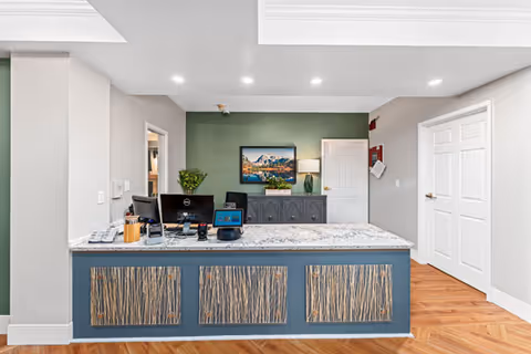 Reception desk area with a granite countertop and blue front panels featuring a natural wood design. Behind the desk is a green accent wall with a landscape painting, a lamp, and a cabinet. The floor is wood with a herringbone pattern, and there are white doors and light gray walls surrounding the area.