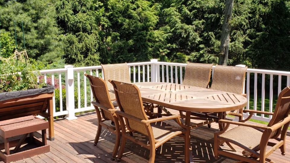 Outdoor wooden deck area with a round wooden table surrounded by six wicker chairs, enclosed by a white railing and surrounded by lush green trees and plants.