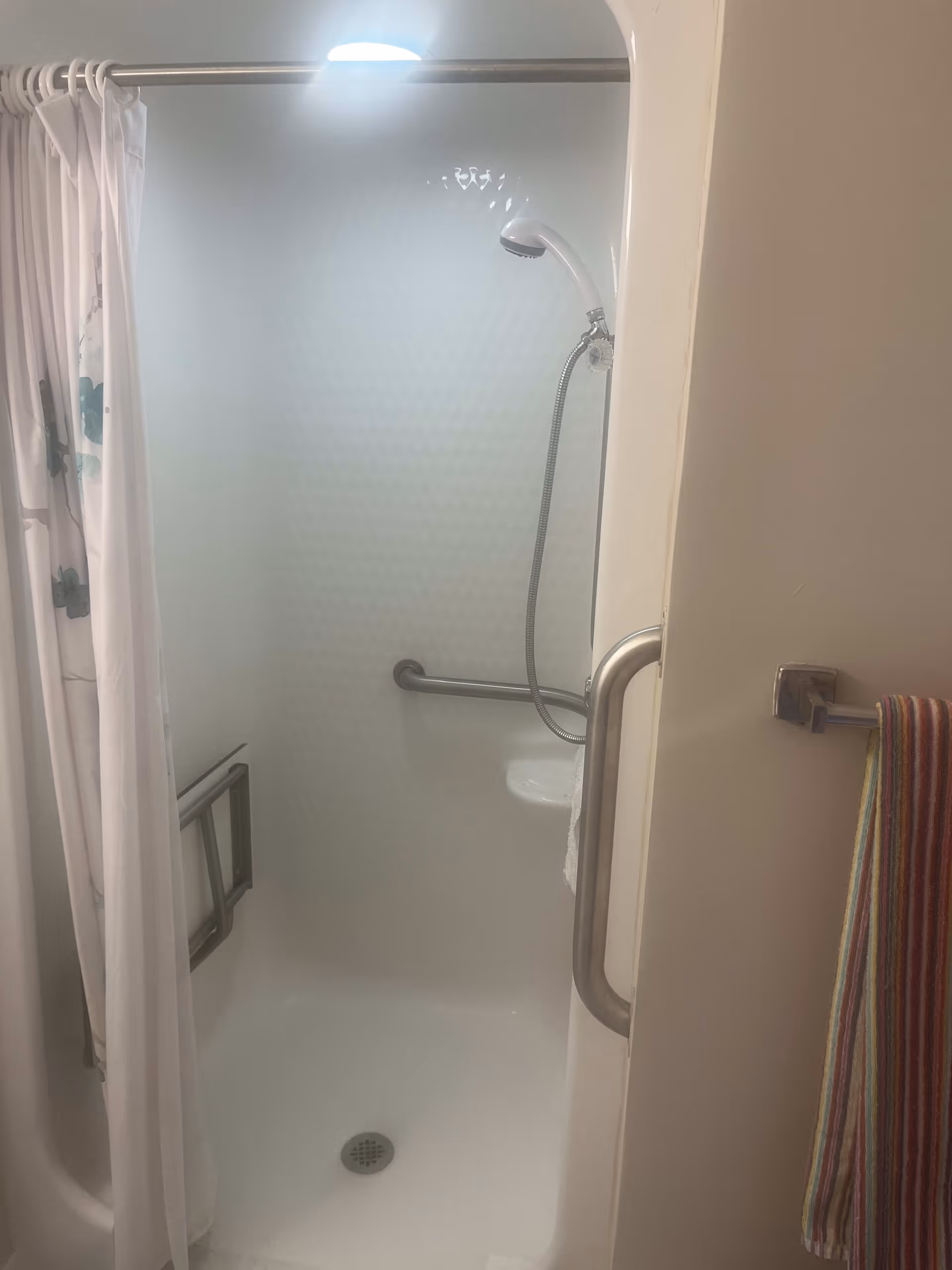 A small shower stall with a white curtain partially drawn to the side. Inside the shower, there is a handheld showerhead attached to a flexible hose, a grab bar on the wall, and a foldable shower seat. A colorful striped towel hangs on a towel rack outside the shower.
