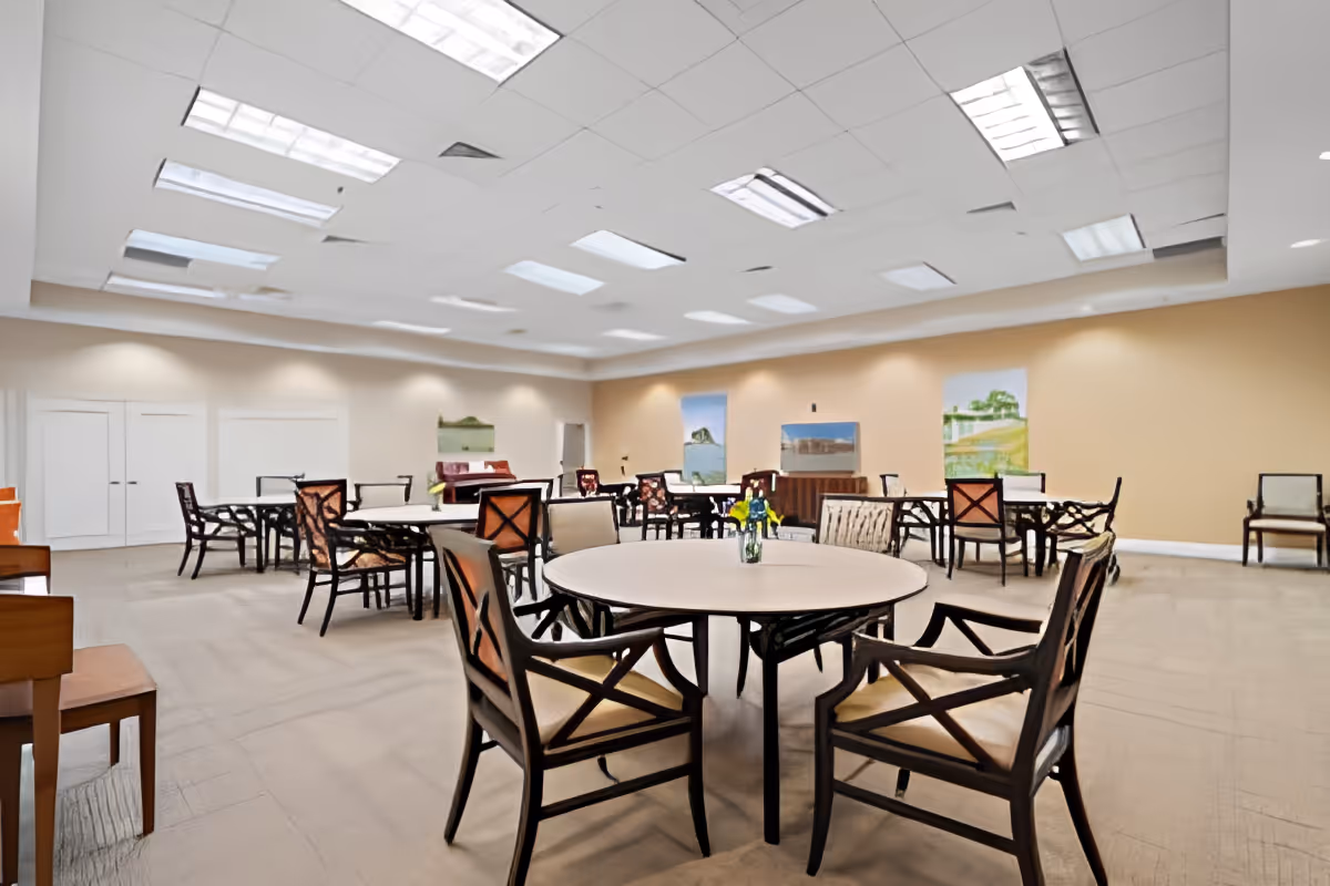Spacious senior community dining/activity room with round tables and chairs under recessed ceiling lights.