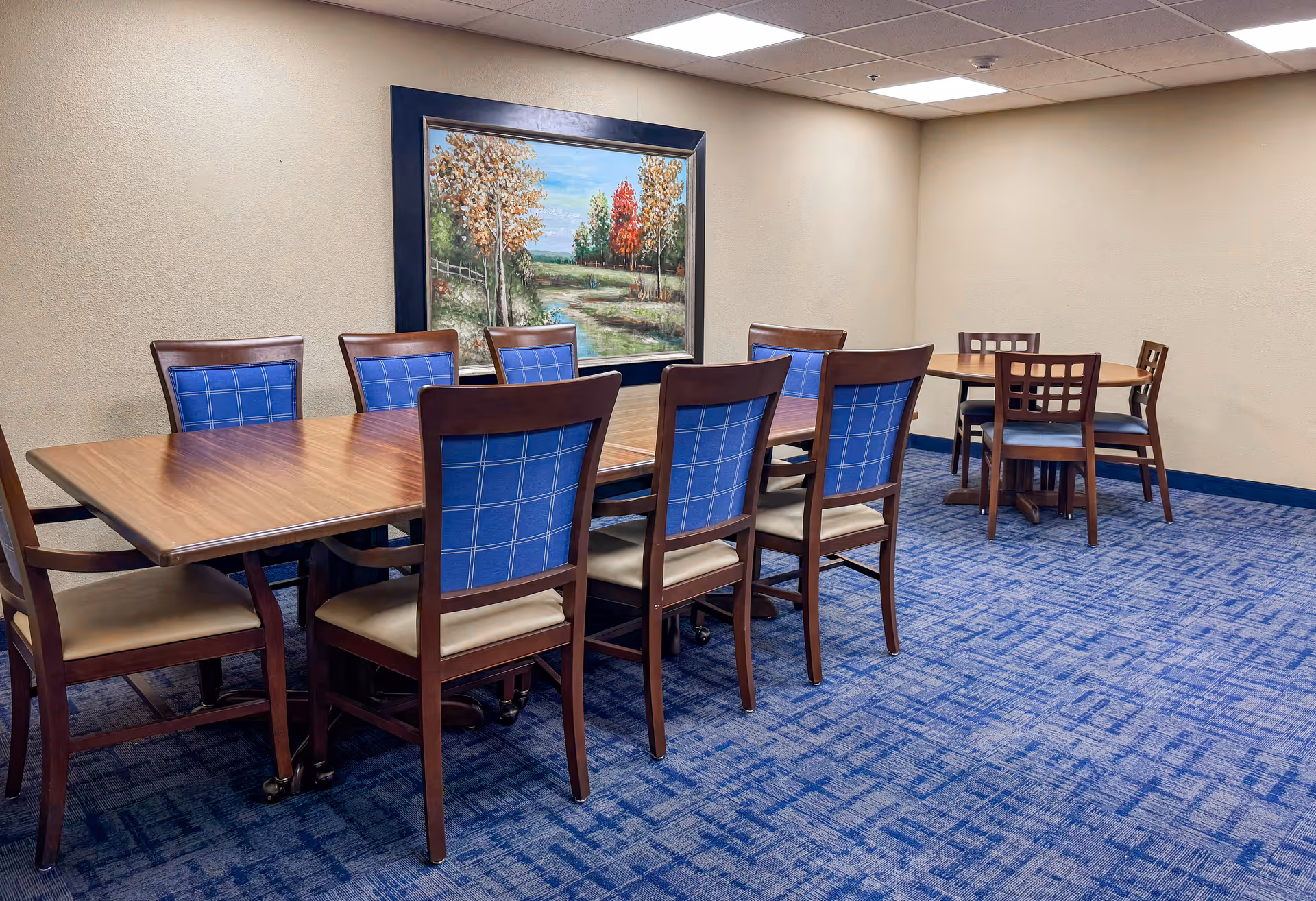 A dining room with two wooden tables and multiple chairs with blue cushioned backs and beige seats. The room has beige walls, a blue patterned carpet, and a large framed painting of a scenic landscape with trees and a path hanging on the wall.