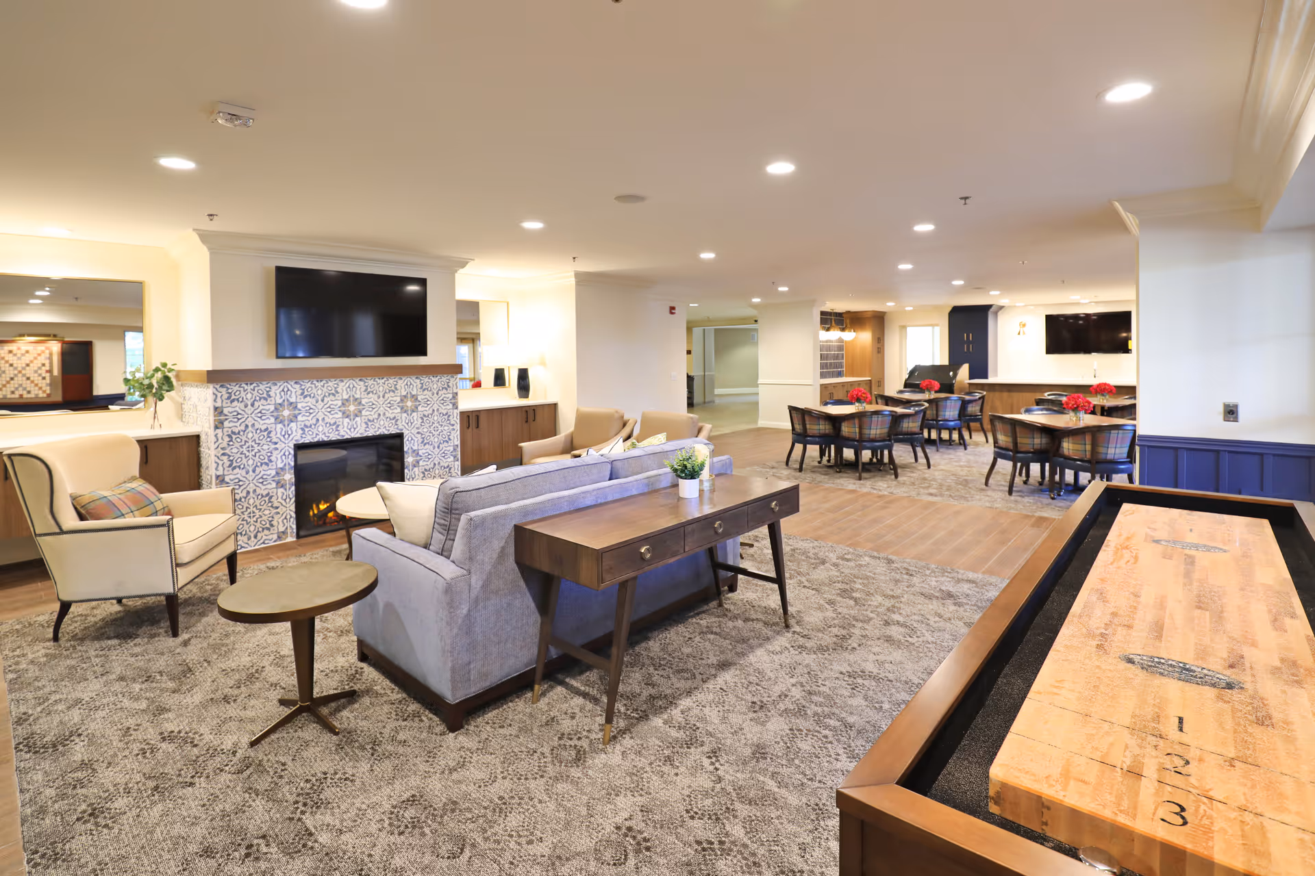 Bright open common room with sofas and chairs around a fireplace and TV, dining tables in the background, and a shuffleboard table in the foreground.
