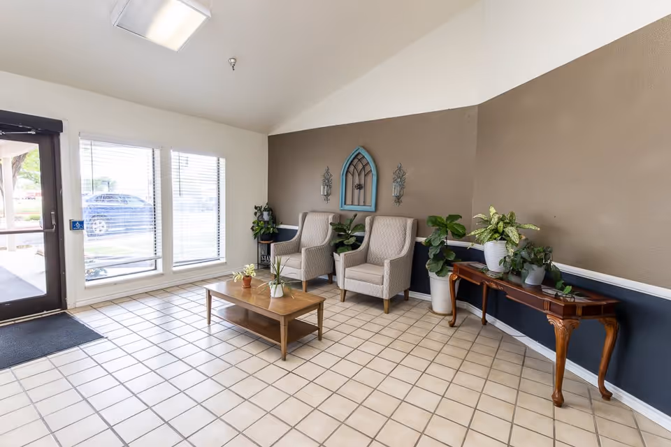A bright and clean lobby area with tiled floor, two beige upholstered armchairs, a wooden coffee table with small potted plants, and a wooden side table with several potted plants. The walls are painted brown and white, with decorative wall sconces and a blue framed mirror. Large windows and a glass door allow natural light to enter the space.