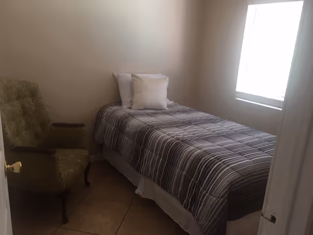 A small bedroom with a single bed covered with a striped gray and white comforter and two white pillows. There is a green upholstered armchair with wooden armrests next to the bed. A window with white blinds lets in natural light on the right side of the room. The floor is tiled and the walls are painted a light beige color.