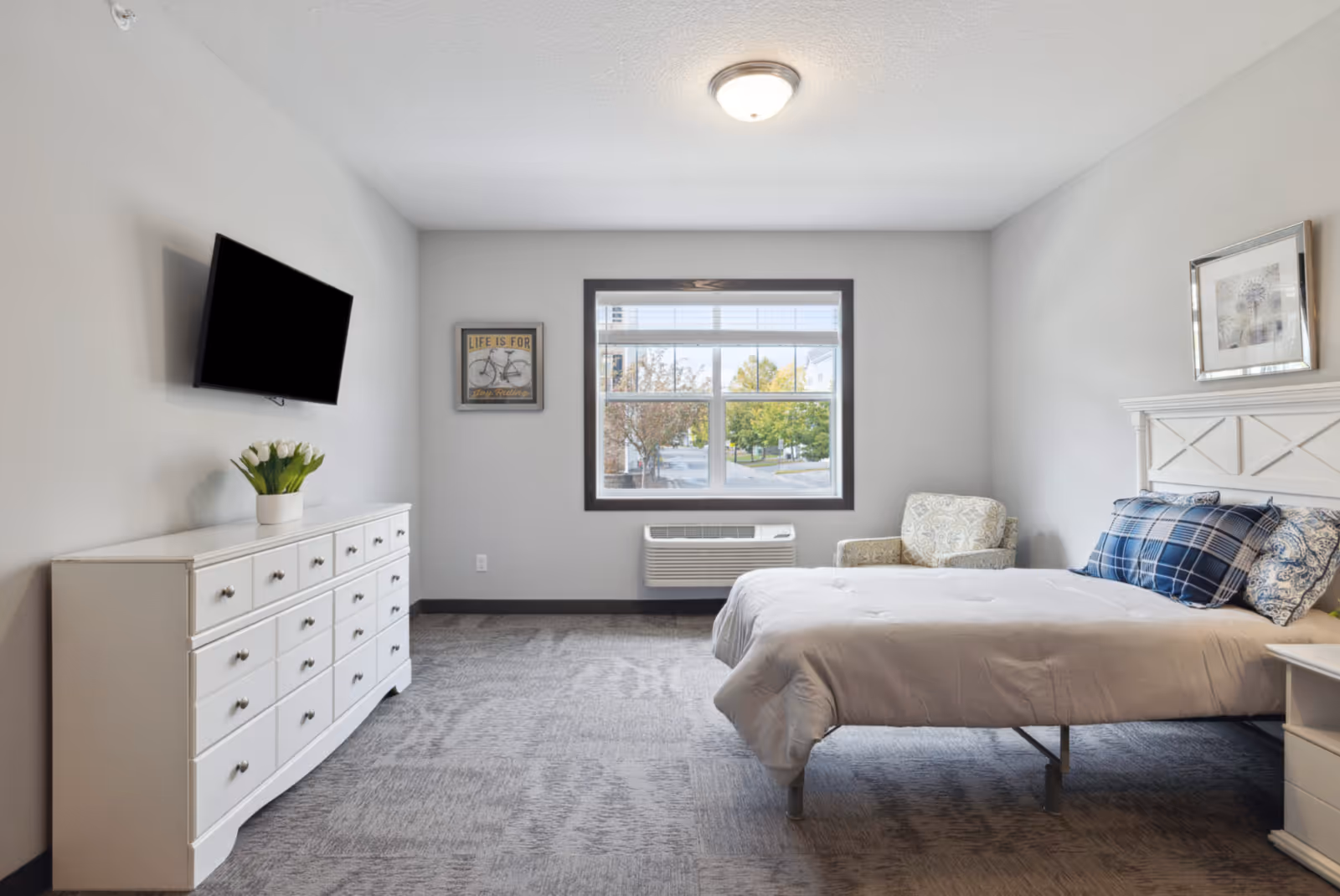 Bright furnished bedroom with a bed, nightstand and armchair, a dresser with a wall-mounted TV, and a window overlooking trees.