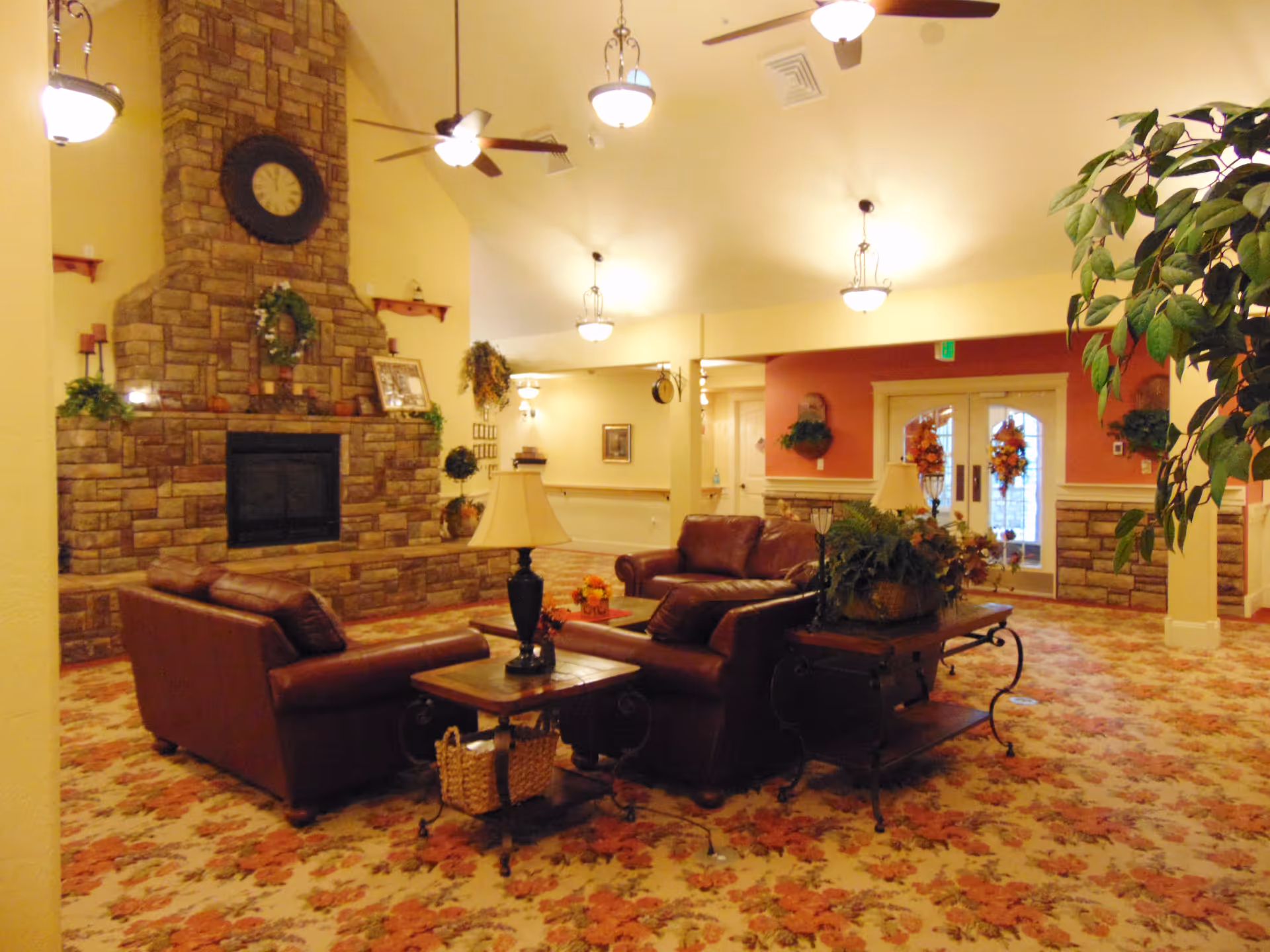 Spacious assisted-living common room with leather sofas and tables arranged around a stone fireplace under pendant lights.