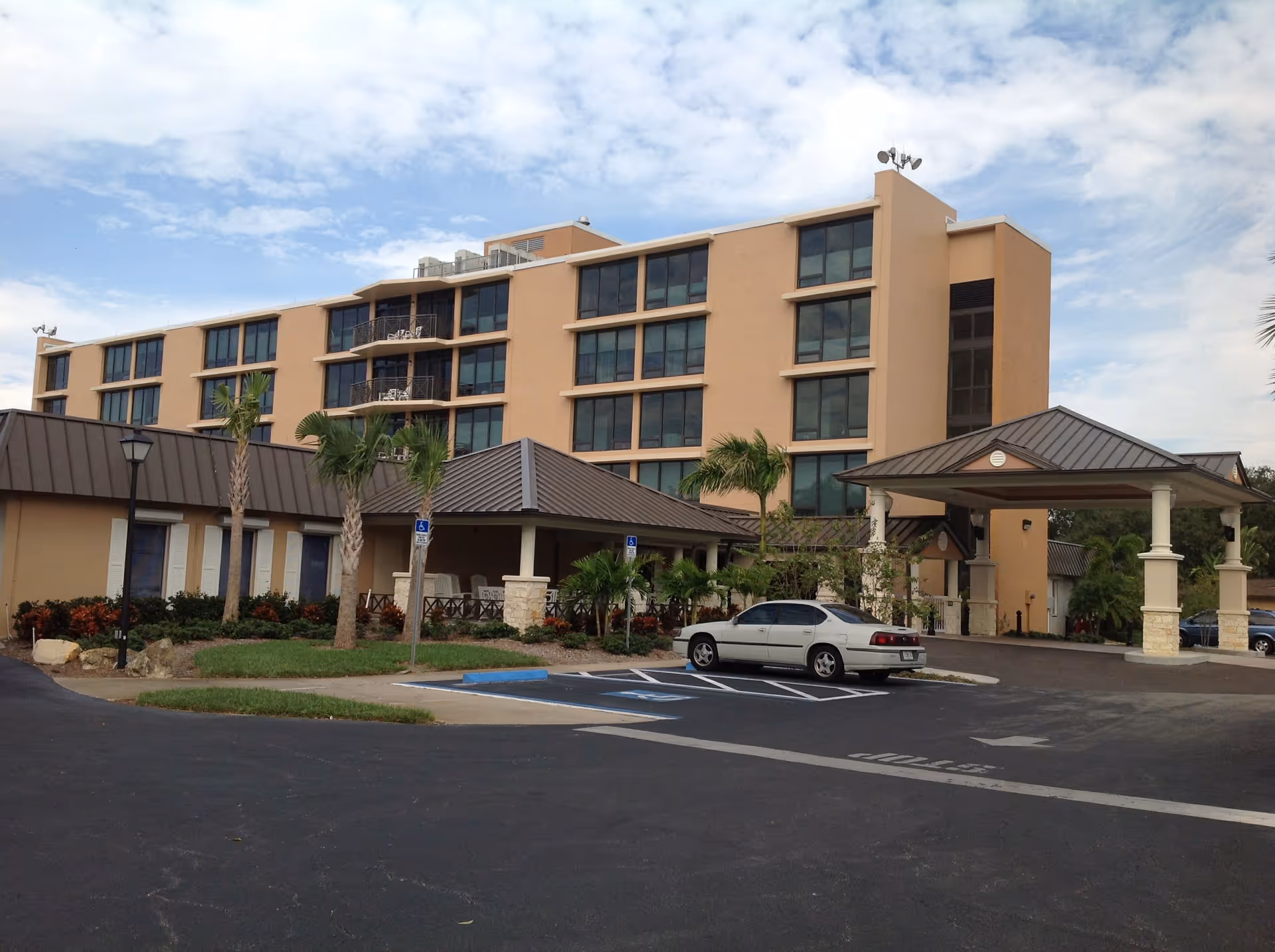 Exterior front of a multi-story beige senior living building with a covered entrance, parking spaces and palm trees.