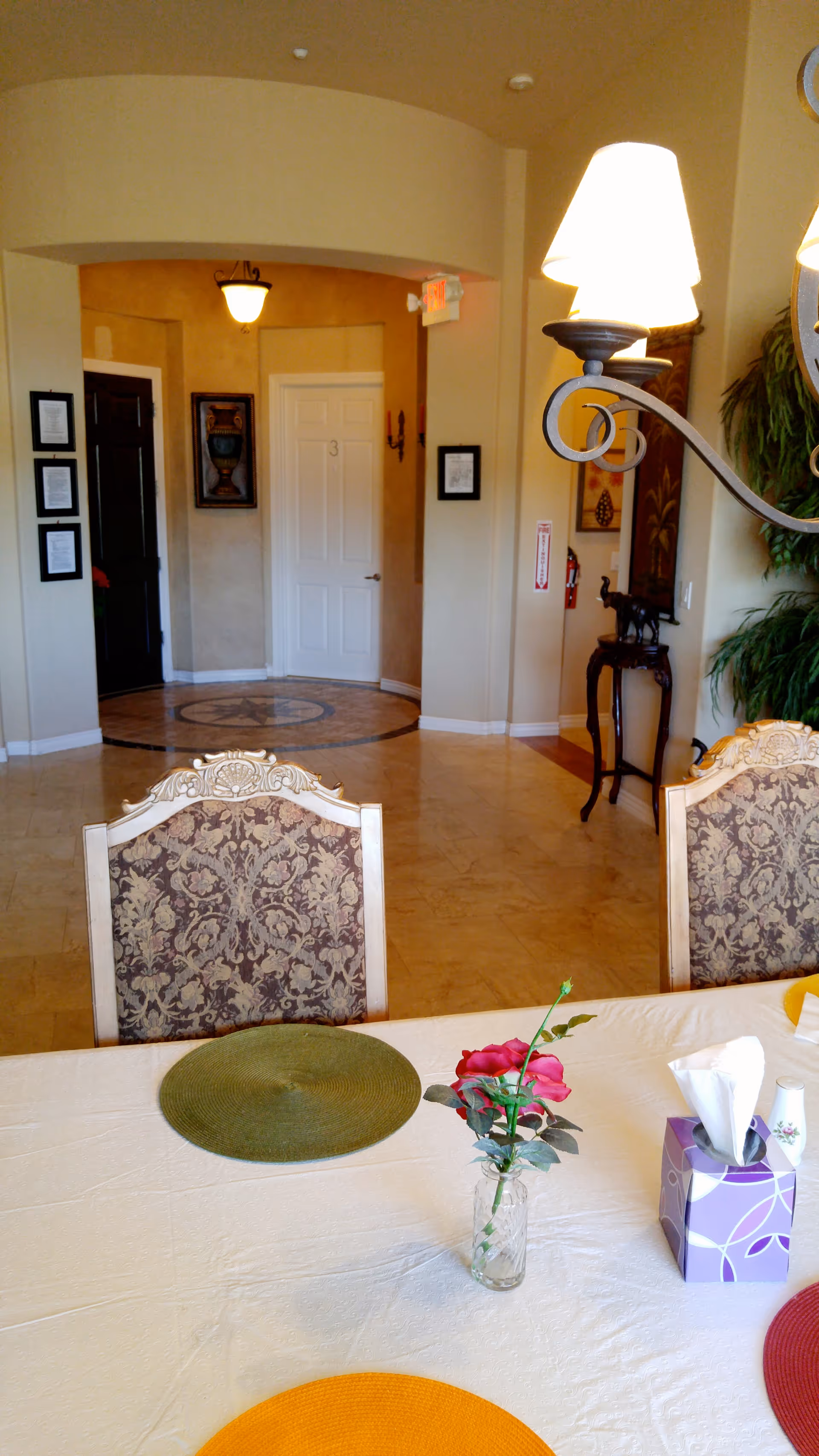View of a dining area in an assisted living facility with a table set with colorful placemats, a small vase with a red rose, and a tissue box. In the background, there is a hallway with a white door numbered 3, framed artwork on the walls, and a decorative floor medallion. The room has warm beige walls and elegant chairs with patterned upholstery.