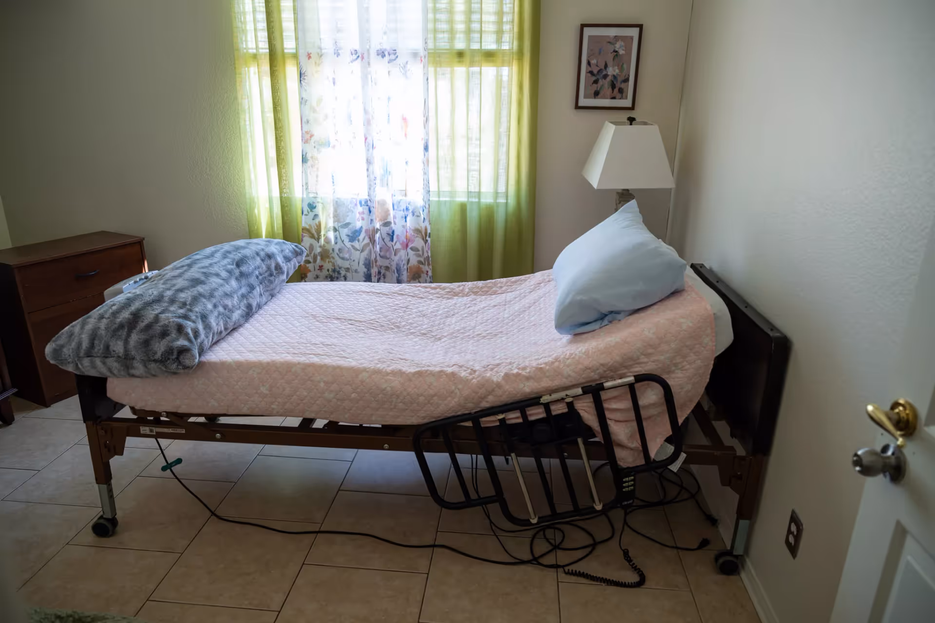 An adjustable hospital bed with pink bedding in a small senior care bedroom.