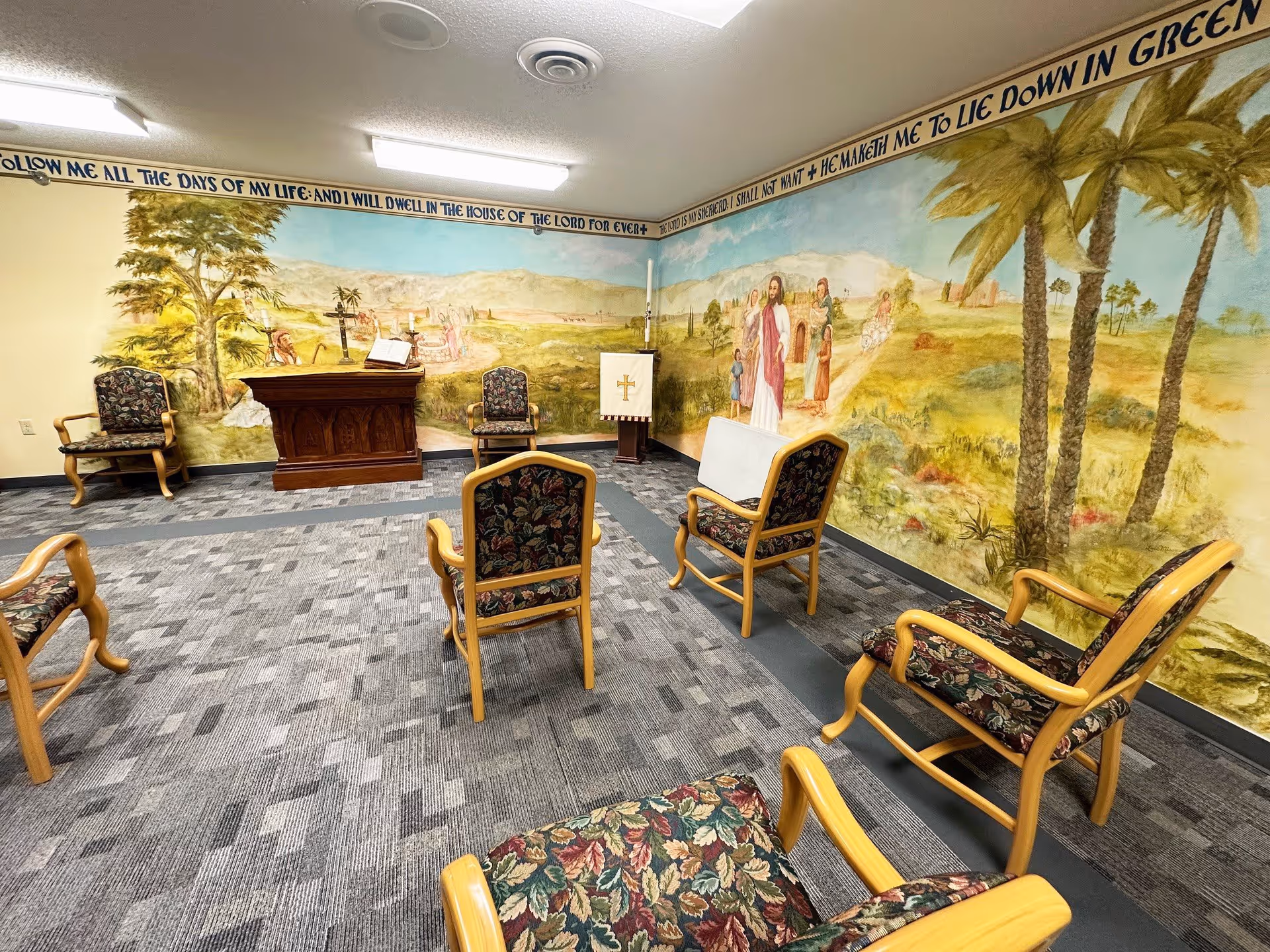 A small chapel or prayer room with floral upholstered wooden chairs arranged in a semicircle facing a wooden altar and a lectern with a cross. The walls feature a large mural depicting a biblical scene with Jesus and several people in a desert landscape with palm trees. Above the mural, a scripture verse is painted around the top edge of the walls. The room has a patterned carpet and fluorescent ceiling lights.