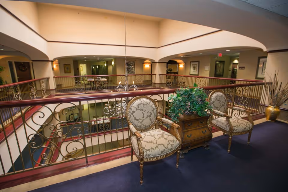 Interior view of a senior living facility showing a second-floor balcony area with ornate railing, two upholstered armchairs with wooden frames, and a small wooden table with a green leafy plant. The area overlooks a lower level with more seating and decorative elements, illuminated by warm lighting.