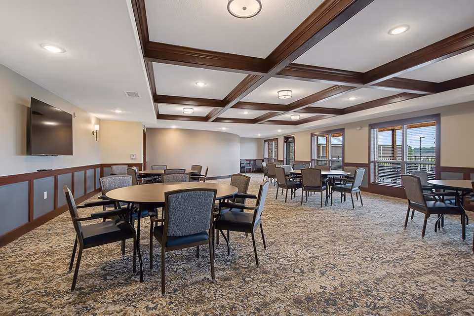 A spacious senior living facility common dining area with multiple round tables and chairs arranged on a patterned carpet. The room features large windows with blinds, a coffered ceiling with recessed lighting, and a wall-mounted flat screen TV.