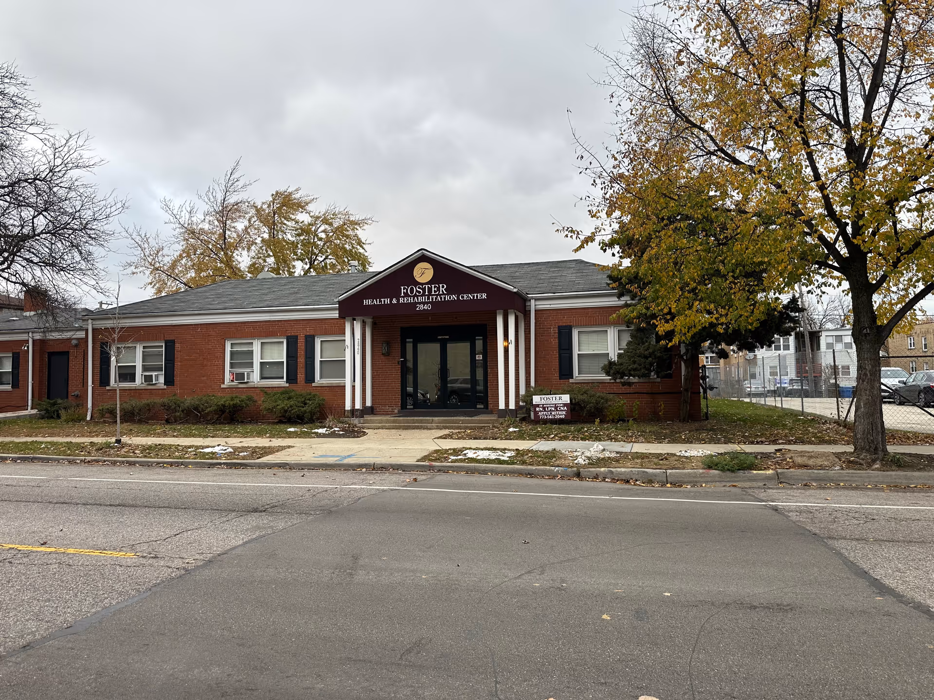 Front view of a single-story brick Foster Health & Rehabilitation Center with a central maroon entrance canopy, sidewalk and trees lining the street.