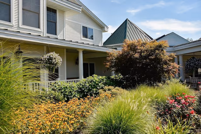 Front exterior of a two-story retirement building with a covered porch and landscaped flower beds and ornamental grasses.