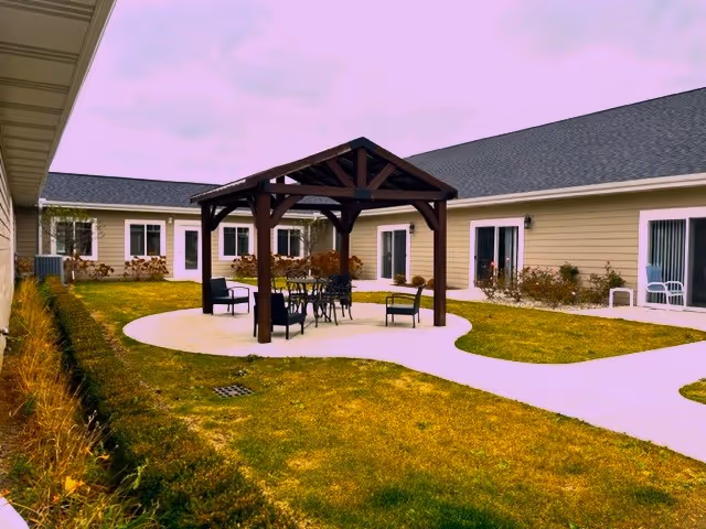 Outdoor courtyard area at Hampton Manor of Trenton Premier Assisted Living Facility featuring a wooden pergola with seating underneath, surrounded by a paved walkway and grass, with the facility building visible in the background.