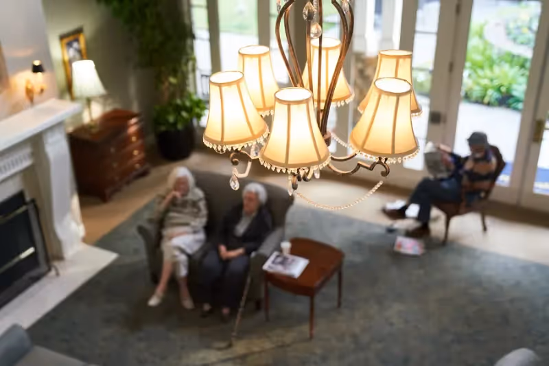 View from above of a cozy living room with a chandelier in the foreground, two elderly women sitting on a couch near a fireplace, and a man sitting on a chair near large glass doors leading outside.