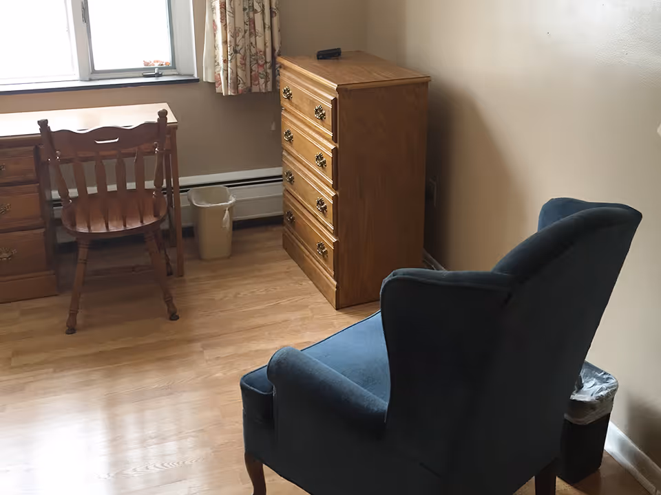 A small room with wooden flooring featuring a wooden desk and chair near a window with floral curtains, a wooden chest of drawers, and a blue upholstered armchair. There are two small trash bins placed near the furniture.