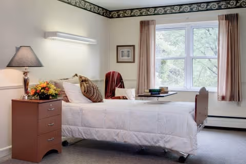 A bright and clean bedroom in a healthcare facility featuring a single bed with white bedding and multiple pillows. Next to the bed is a wooden nightstand with a lamp and a floral arrangement. A chair with a red throw is positioned near a window with beige curtains, allowing natural light to fill the room.