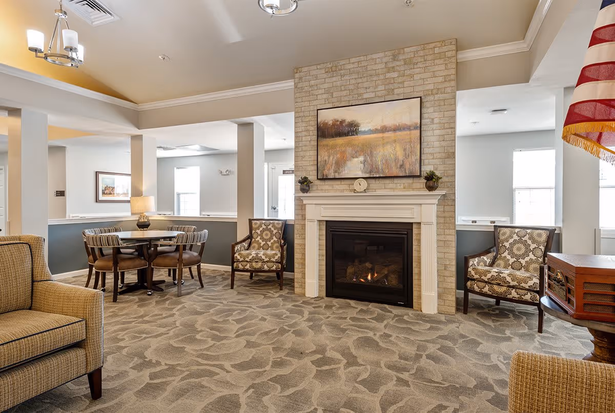 A cozy senior living common area with a lit fireplace set in a brick wall, a landscape painting above the mantel, two patterned armchairs on either side, a round table with four chairs, and beige patterned carpet. The room is well-lit with natural light from windows and ceiling lights.