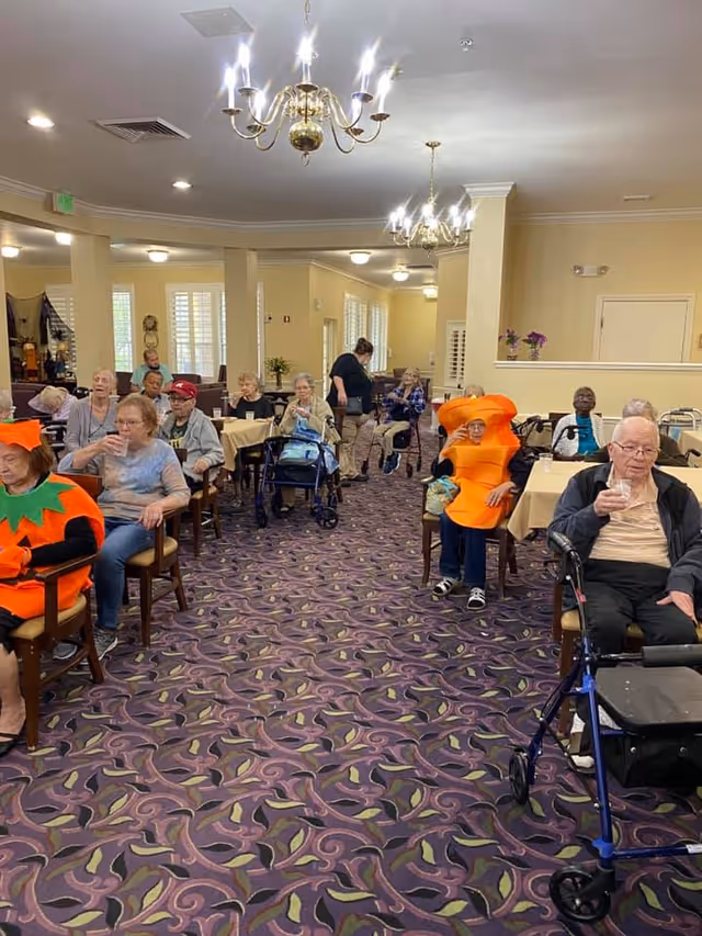 A group of elderly people sitting at tables in a decorated dining room or common area, some wearing festive costumes including a pumpkin and carrot outfit. The room has patterned carpet, chandeliers, and beige walls with windows and plants in the background.