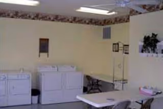 Laundry room with two washing machines and a dryer against a wall, a table with chairs, and a small desk with a chair in the corner. The walls are light-colored with a floral border near the ceiling.
