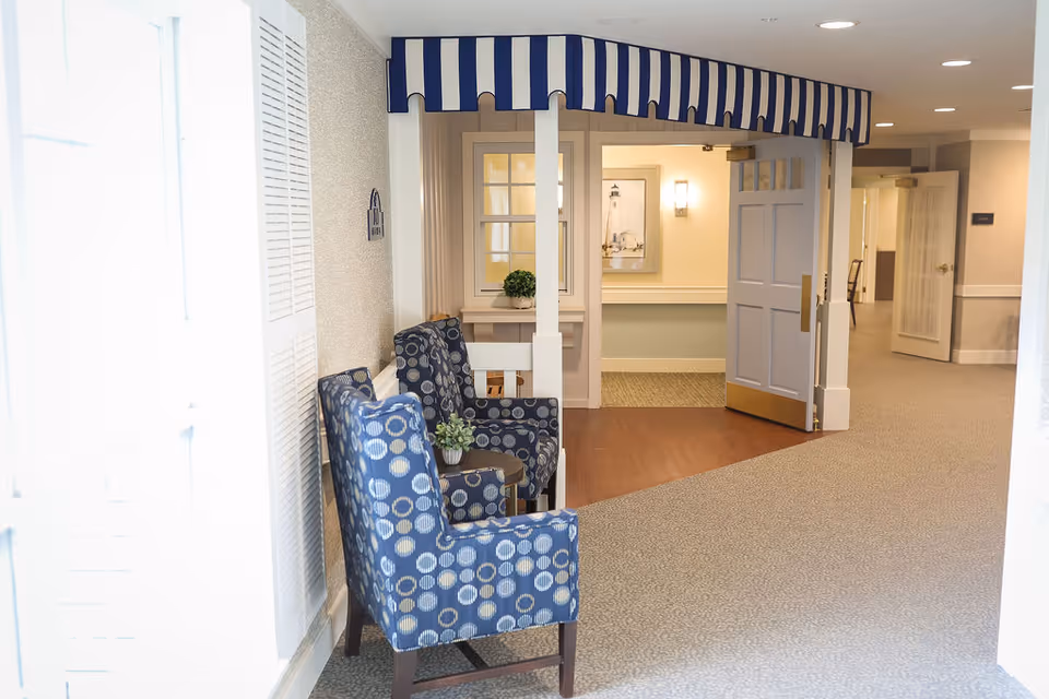 A cozy indoor seating area with two blue patterned armchairs and a small table with a plant between them. The area is decorated with a blue and white striped canopy overhead. The background shows an open door leading to a hallway with soft lighting and framed artwork on the wall.