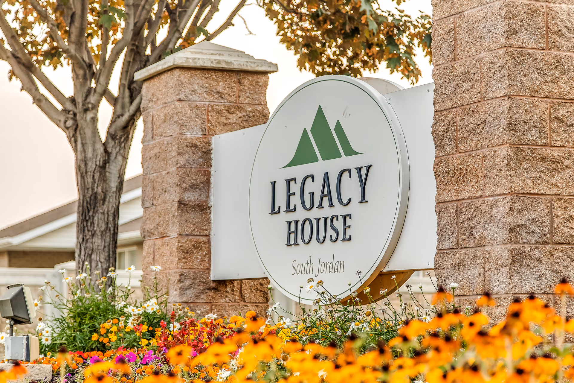 A large round sign mounted between two stone pillars reads 'Legacy House South Jordan' with a green mountain logo above the text. The sign is surrounded by colorful flowers including orange, yellow, white, and purple blooms. A tree and part of a building are visible in the background.