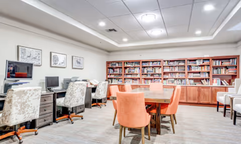 A well-lit room with a large wooden table surrounded by orange upholstered chairs in the center. Along one wall, there is a built-in wooden bookshelf filled with books. On the opposite side, there are desks with computers and office chairs. The walls are decorated with framed artwork, and the ceiling has recessed lighting.