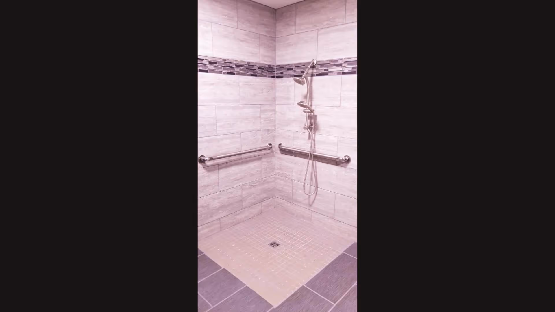 A tiled walk-in shower with two stainless steel grab bars on adjacent walls and a dual showerhead fixture mounted on the wall. The shower walls have light-colored tiles with a decorative horizontal strip of smaller, darker tiles near the top.