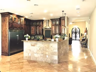 A spacious kitchen with dark wooden cabinets, a large stainless steel refrigerator, a central island with a stone facade, and decorative plants on the countertop. The kitchen opens into a hallway with an arched doorway and a decorative wrought iron door at the end.