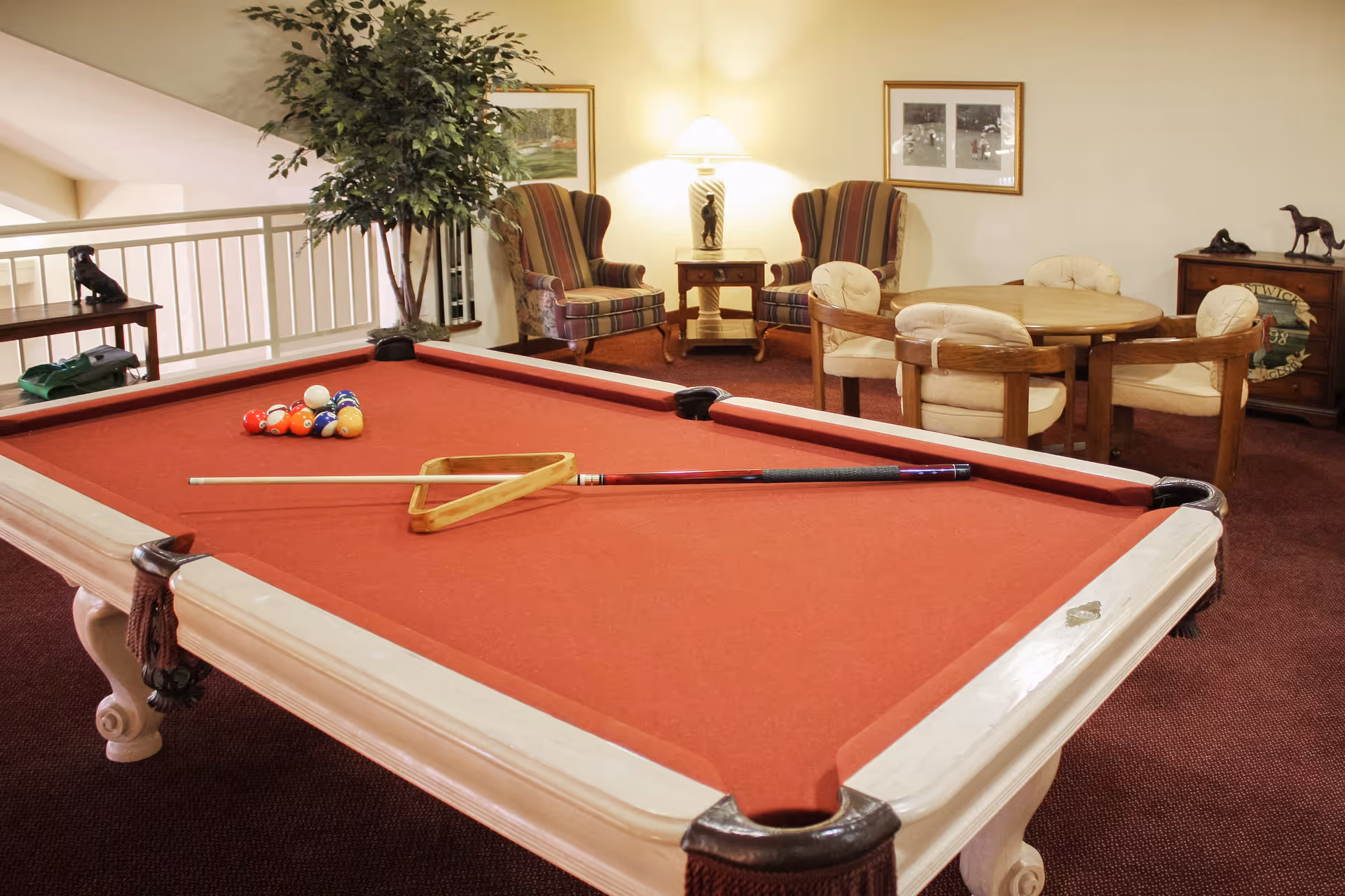 A recreational room featuring a red-felt pool table with a cue stick and triangle rack on it. Behind the pool table, there are two striped armchairs with a small table and lamp between them. To the right, there is a round wooden table with four cushioned chairs. The room has carpeted flooring and framed pictures on the walls, along with decorative items on side tables.