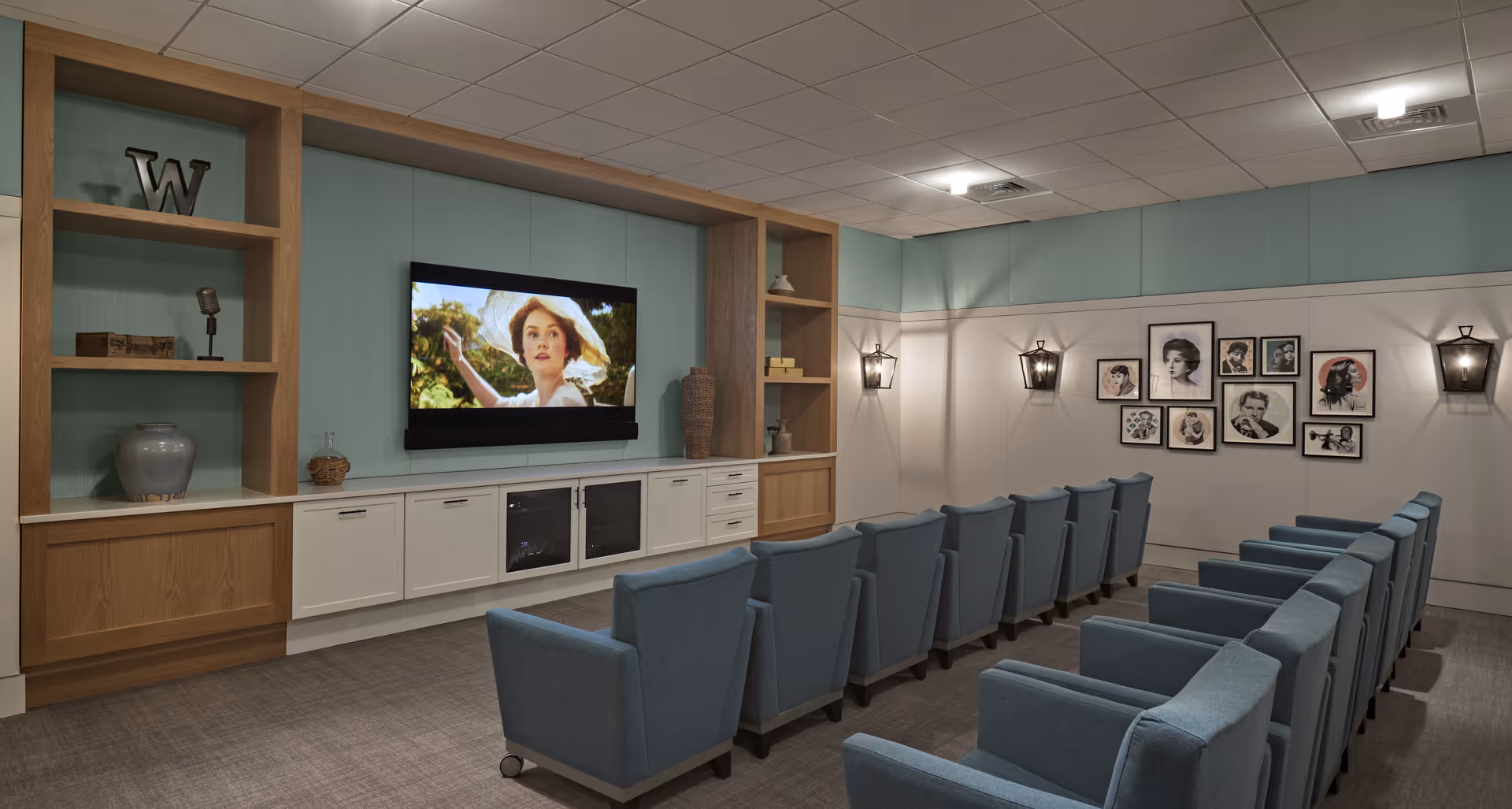 A cozy media room with two rows of blue upholstered armchairs facing a wall-mounted flat screen TV. The TV is playing a scene featuring a woman in a white hat. The room has built-in wooden shelves and cabinets painted white, with decorative items such as vases and a letter 'W'. The walls are light blue and white, adorned with framed black and white portraits and wall-mounted lantern-style lights.