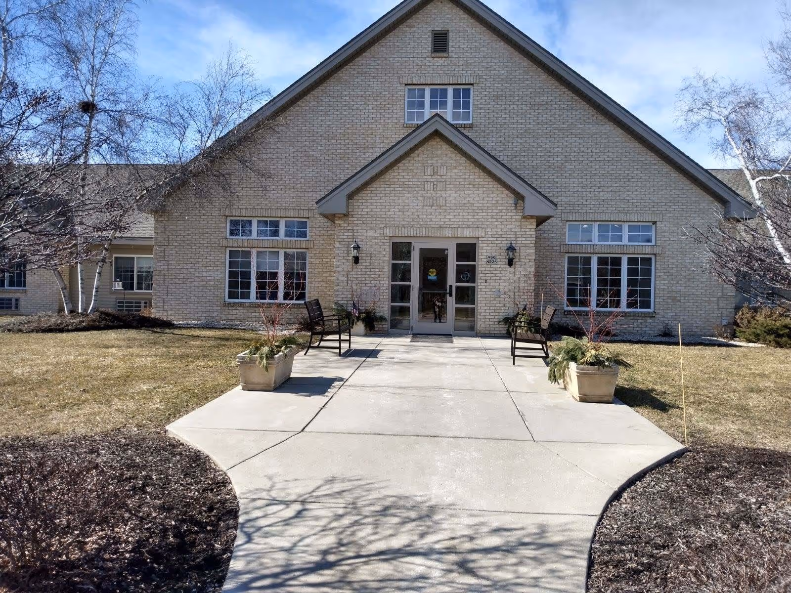 Front exterior view of a single-story brick building with a peaked roof, large windows, and a glass door entrance. There are two benches and potted plants on either side of the concrete walkway leading to the entrance. Leafless trees and a clear blue sky are visible in the background.