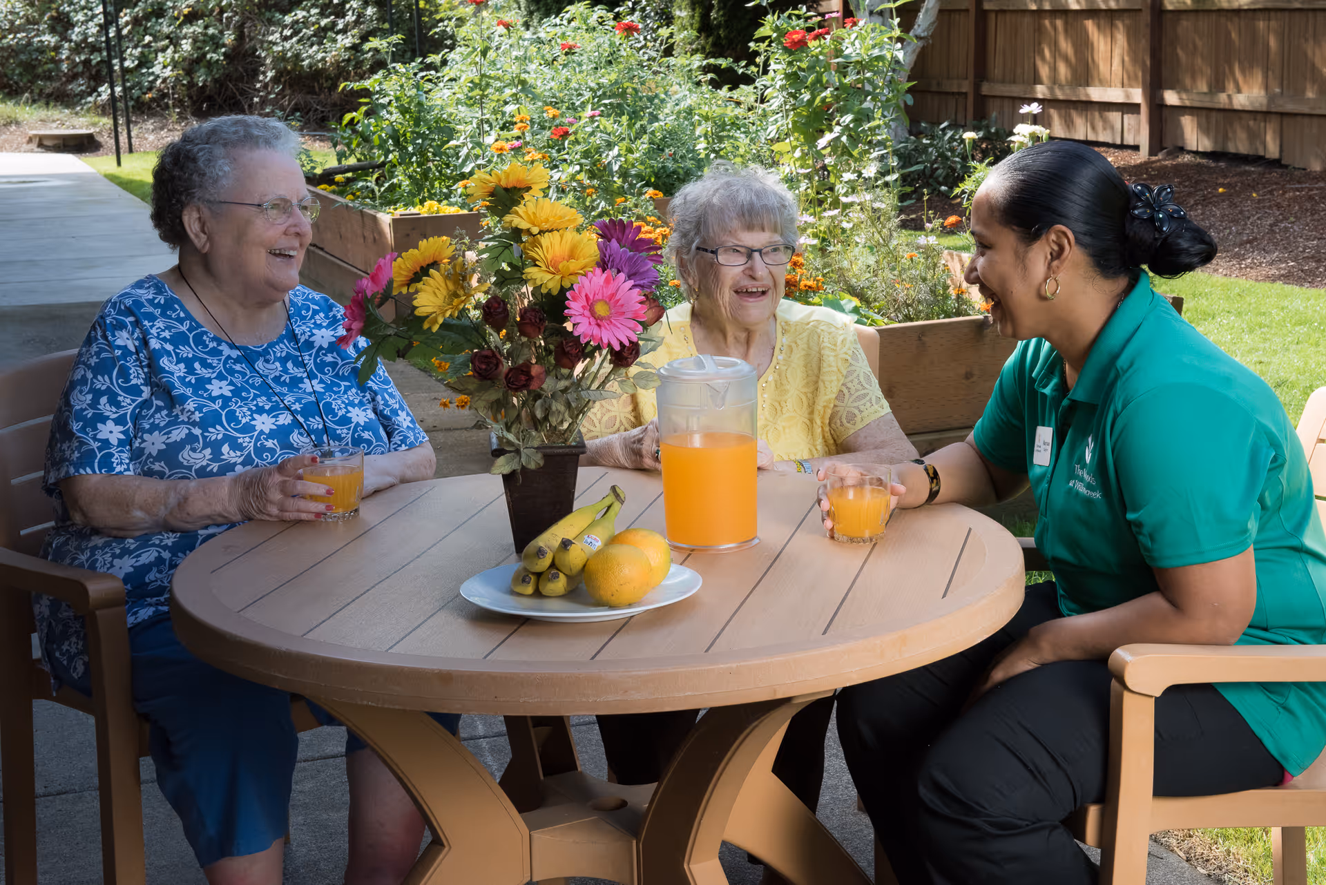 Two elderly women and a caregiver sitting around a round outdoor table with a pitcher of orange juice, glasses, a plate of bananas and oranges, and a vase of colorful flowers, enjoying a conversation in a garden setting.