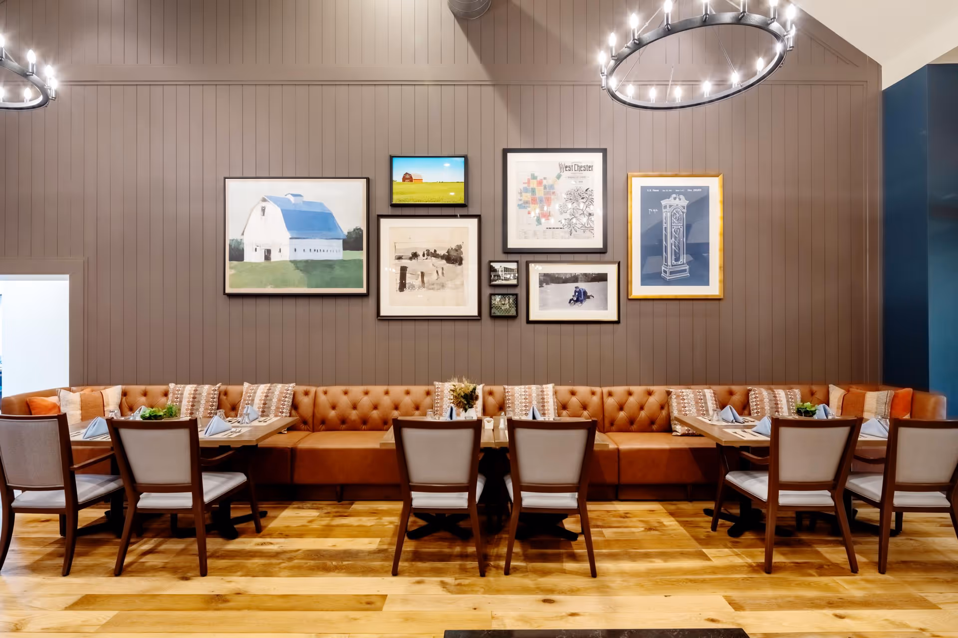A cozy dining area with a long brown tufted bench along a paneled wall adorned with various framed artworks and pictures. There are three wooden tables set with napkins and small plants, each surrounded by beige cushioned chairs. Two circular chandeliers hang from the ceiling, illuminating the space with warm light. The floor is made of light wood planks.