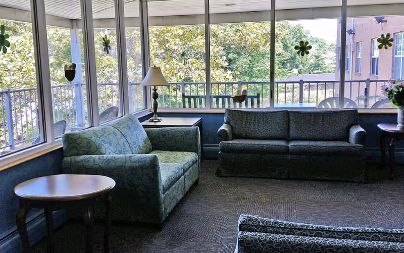 A sunlit sitting area with two patterned sofas, a round wooden side table, a lamp on a small table, and large windows overlooking green trees and part of a building outside.