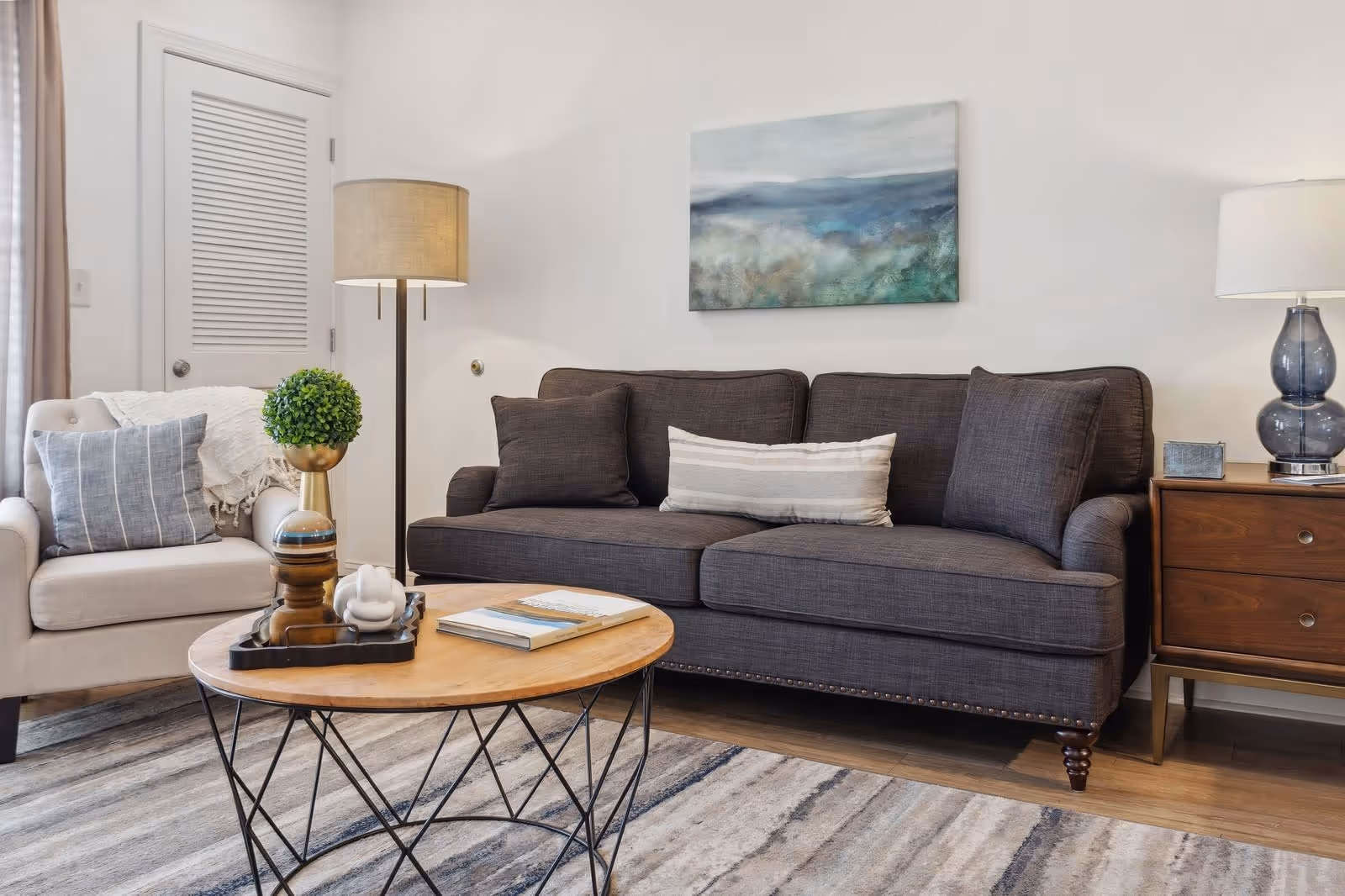A cozy living room with a dark gray sofa adorned with three pillows, a beige armchair with a striped pillow and a white throw, a round wooden coffee table with decorative items and books, a floor lamp, a wooden side table with a blue glass lamp, and a landscape painting on the white wall.