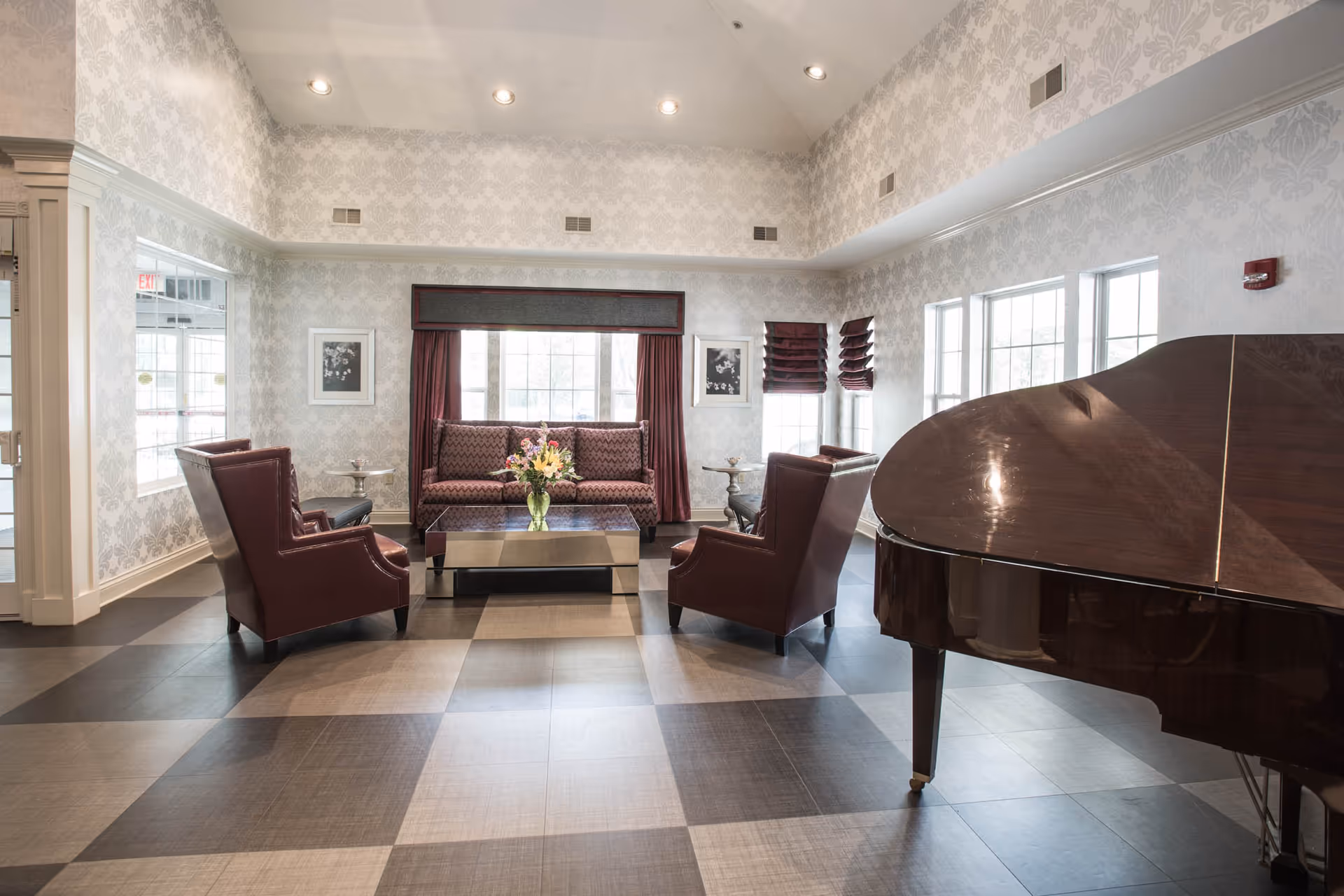 Bright common room with high ceilings featuring a grand piano, leather armchairs, a sofa, coffee table with flowers, and large windows.