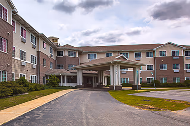 Exterior view of a three-story senior living facility building with a covered entrance driveway, surrounded by a small lawn and paved parking area under a cloudy sky.