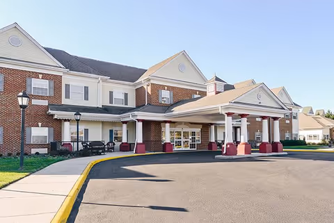 Exterior view of a senior living facility building with a covered entrance supported by white columns with red bases, brick and beige siding walls, and multiple windows. There is a paved driveway and sidewalk with street lamps on a sunny day.