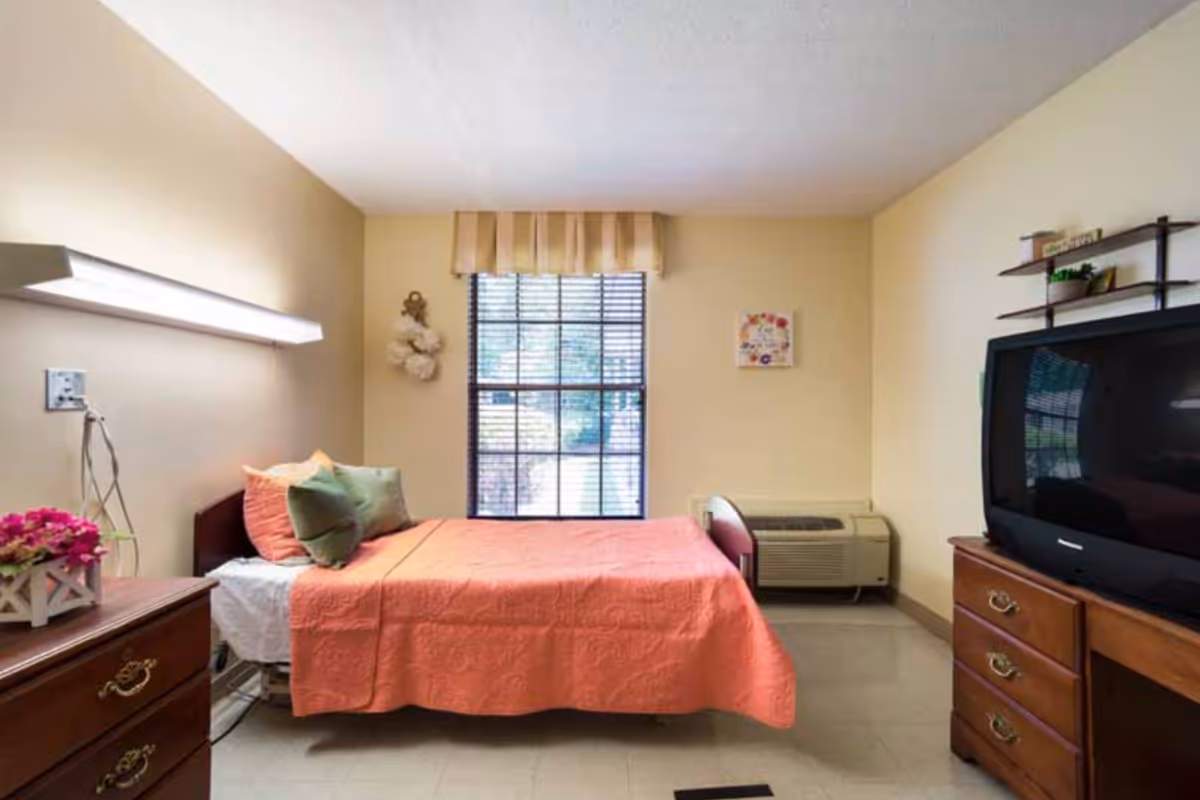 Small furnished bedroom with a single bed covered in a coral bedspread, window blinds, dressers, and a TV.